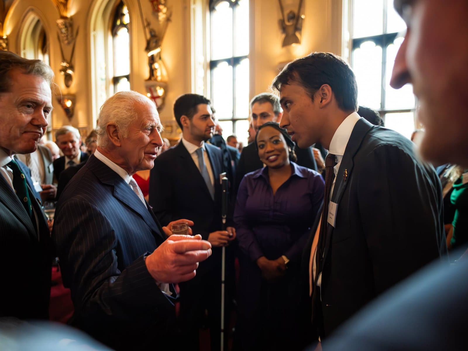King Charles III, glass in hand, in conversation with Sunny Jain among guests at a reception in a grand hall